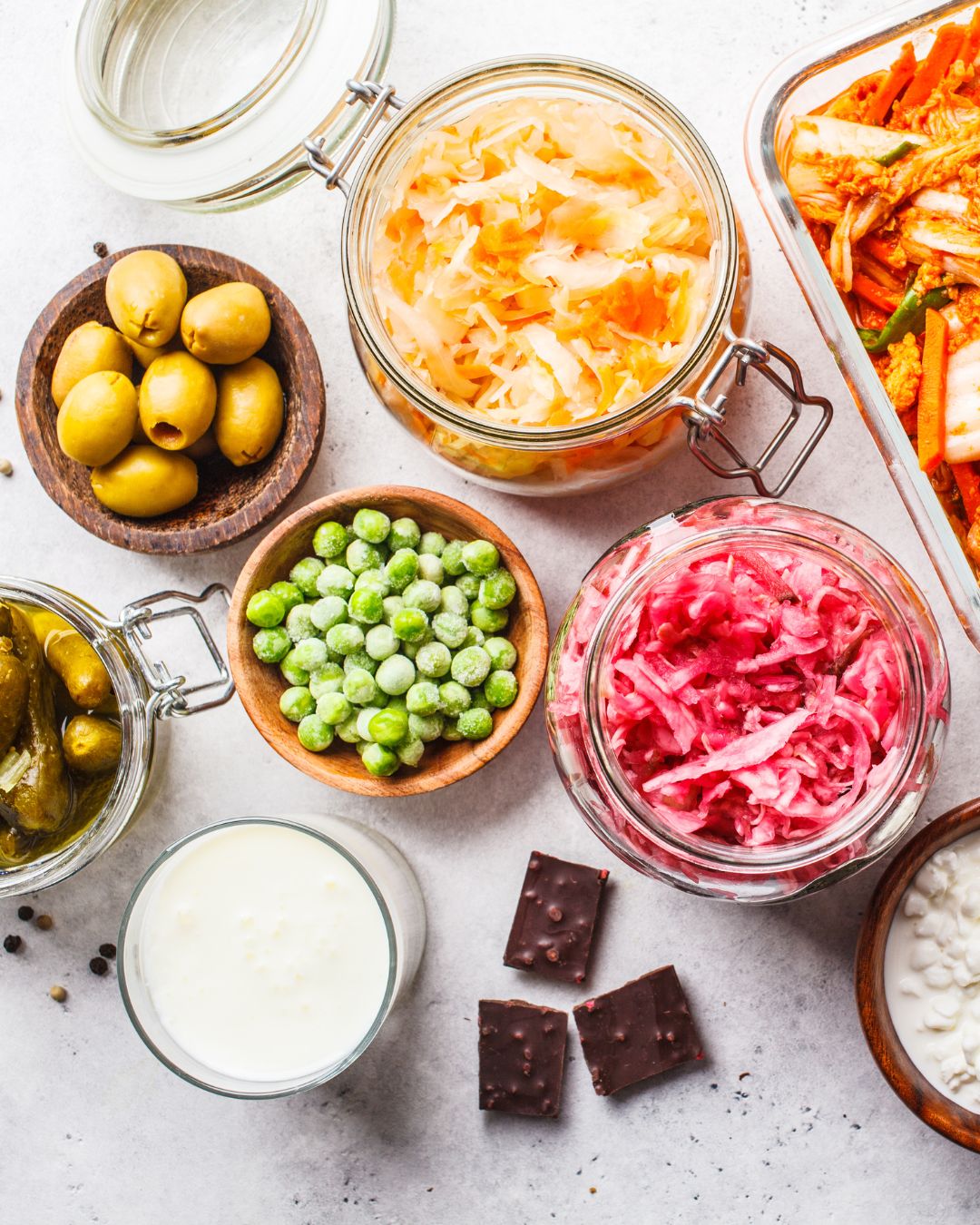 A variety of fermented foods displayed on a white background. The image features a glass jar of sauerkraut, a dish of kimchi, green olives in a wooden bowl, frozen green peas in another wooden bowl, a glass of milk kefir, pickles in a jar, dark chocolate squares, and a jar of red fermented vegetables, possibly beet kraut. The arrangement showcases different types of fermented foods and beverages, along with a few non-fermented items like peas and chocolate.